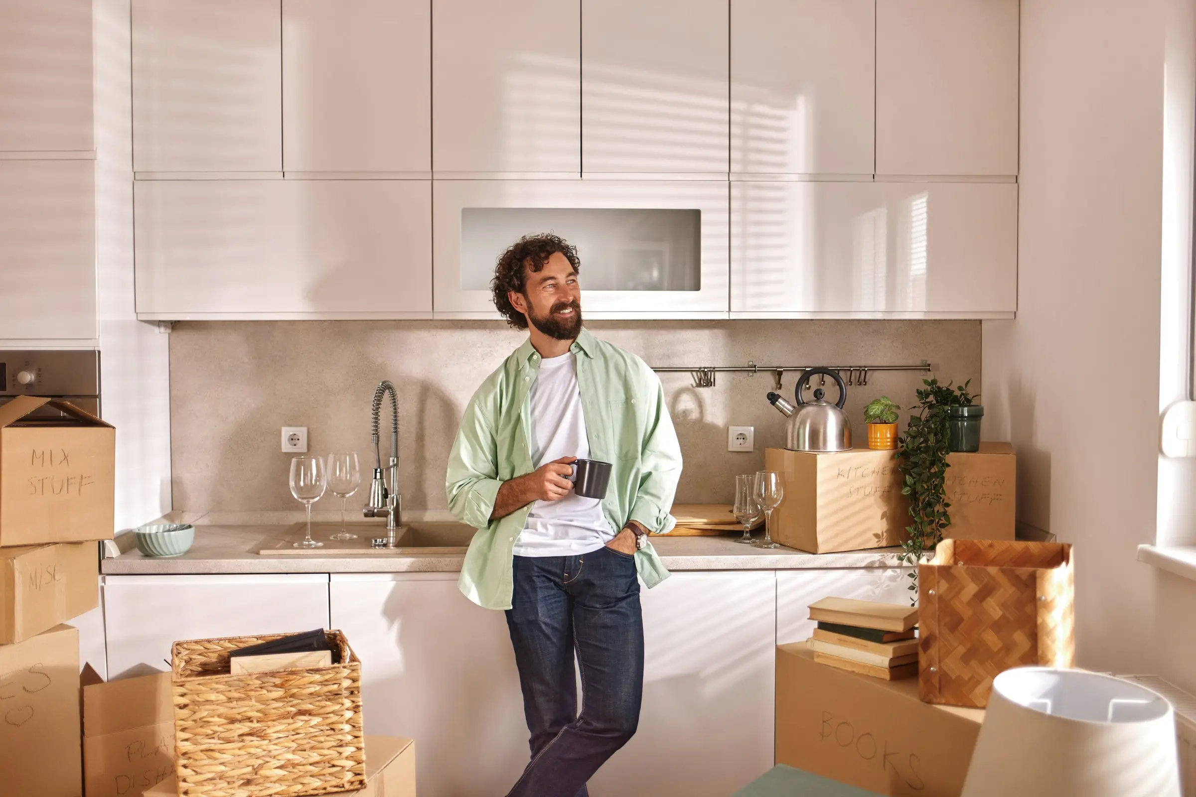 Happy homeowner enjoys a cup of coffee in his new modern kitchen, surrounded by unpacked boxes, feeling relaxed and satisfied after moving in